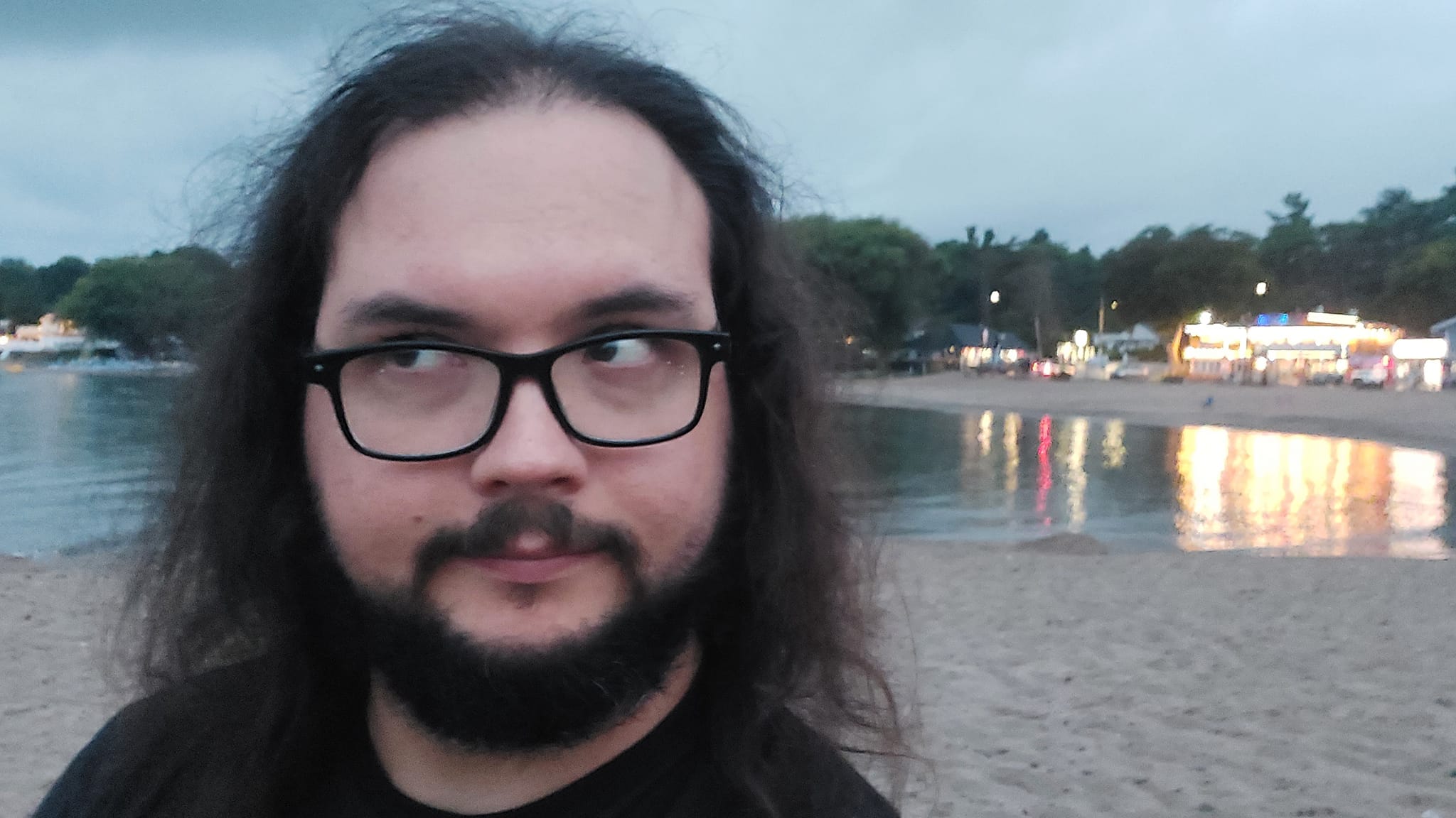 A man with long dark hair and a beard is at a sandy beach with a granite boulder breakwater, photo 5